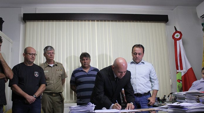 Homem careca se abaixa para assinar um papel sobre uma mesa, circundado por outros homens de pé com uma cortina ao fundo, em uma sala de gabinete