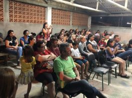 Mães e pais de alunos sentados em carteiras velhas em uma espécie de galpão, durante uma reunião, todos olham para o mesmo lado