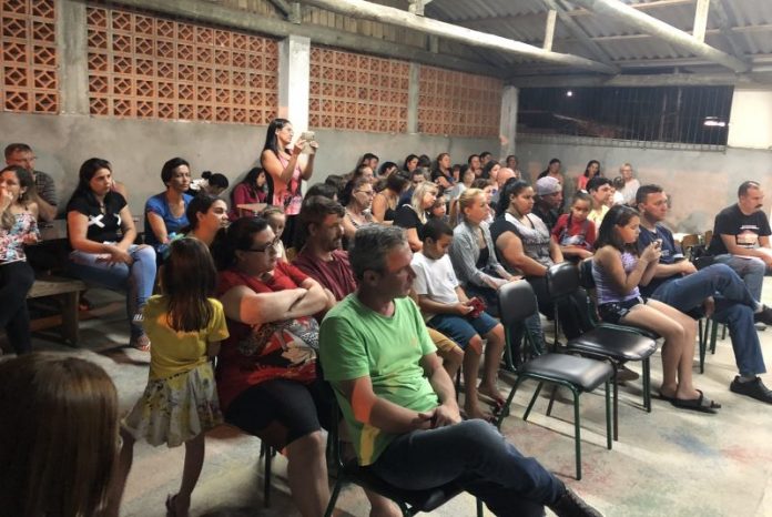 Mães e pais de alunos sentados em carteiras velhas em uma espécie de galpão, durante uma reunião, todos olham para o mesmo lado