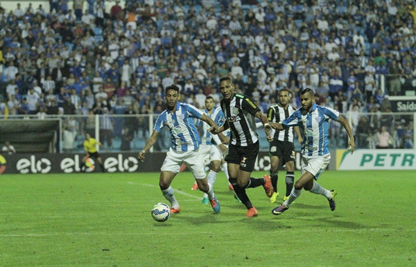 Jogadores de figueirense e avaí, com camisas listradas, disputam a bola no gramado, com a torcida observando ao fundo a partida noturna