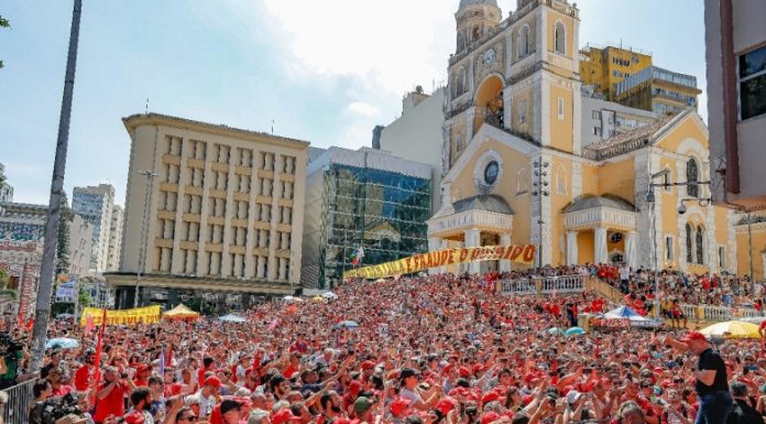 Multidão em frente à Catedral de Florianópolis vê Lula discursar, usando camiseta preta e boné vermelho do MST