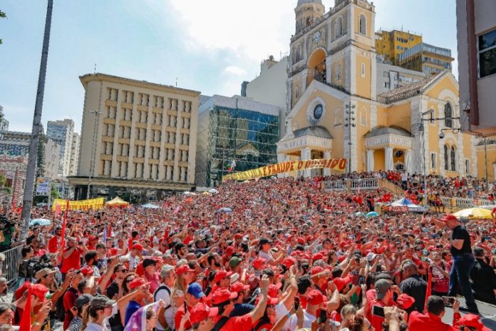 Multidão em frente à Catedral de Florianópolis vê Lula discursar, usando camiseta preta e boné vermelho do MST