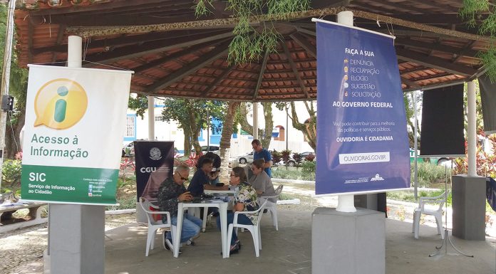 Coreto da praça com banners pendurados, com pessoas conversando em mesas e cadeiras