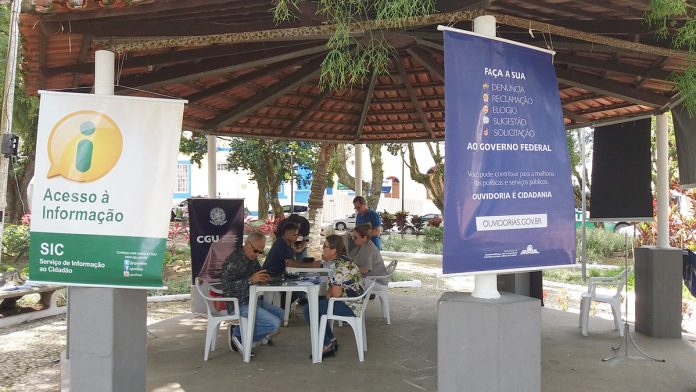 Coreto da praça com banners pendurados, com pessoas conversando em mesas e cadeiras