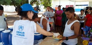 Mulher vista de costas, usando camiseta branca da ACNUR e boné azul entrega refeição a um homem, com um uma fila de venezuelanos atrás