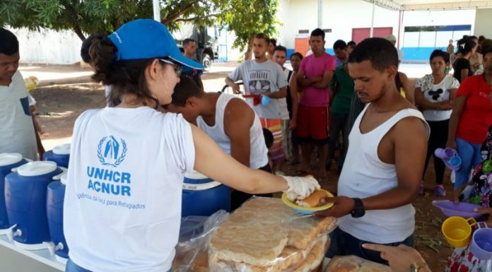 Mulher vista de costas, usando camiseta branca da ACNUR e boné azul entrega refeição a um homem, com um uma fila de venezuelanos atrás