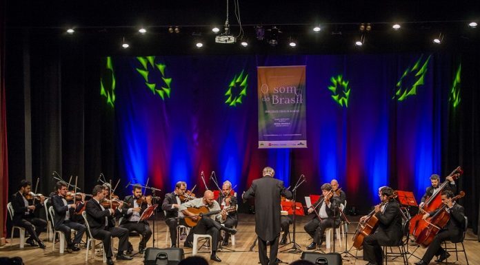 Orquestra se apresenta em um palco, com o maestro à frente, iluminada por uma luz azul de fundo