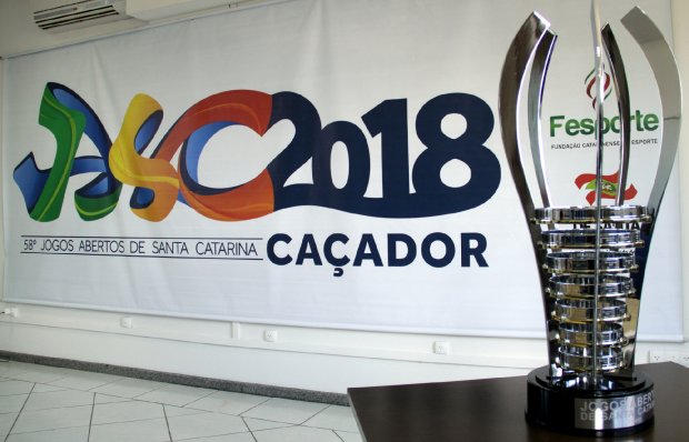 Imagem do troféu de campeão geral em primeiro plano, à direito, e ao fundo o cartaz oficial com os dizeres 