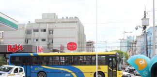 Vista lateral de um ônibus coletivo azul e amarelo cruzando uma avenida com um calçadão no meio