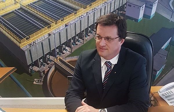 Zanotto sentado em uma mesa usando terno preto e óculo à frente de um painel com fotos da Casan