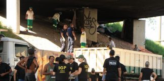 Foto em baixo do viaduto mostrando agentes de segurança na rua falando com usuários de drogas; ao fundo funcionários limpam e jogam lixo em caminhão