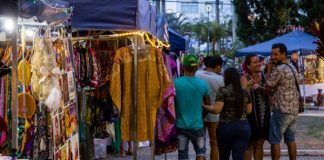 pessoas passam em frente à barracas de feiras com roupas à venda em uma praça no final do entardecer