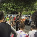 Garota negra lê uma poesia em seu celular de pé no centro de uma roda de espectadores numa praça