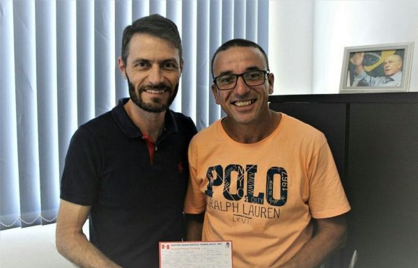 Dois homens posam para foto segurando a ficha de filiação de reni (à direita), usando camiseta laranja óculos; os dois estão no gabinete