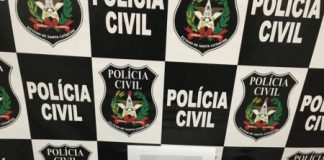 Mesa com armas, celulares e munições com um painel de logos da polícia civil ao fundo