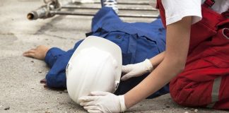 Operário de construção civil usando macacão azul e capacete branco estendido no chão sendo atendido por uma mulher de macacão vermelho e luvas brancas