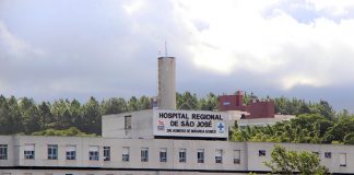 Vista externa e distante da fachada e parte superior do hospital, onde se lê o nome na parte mais alta do prédio, com árvores na frente e atrás