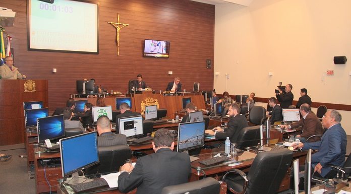 Salão da câmara de vereadores onde se vê ao fundo à esquerda a tribuna com um vereador falando e demais sentados em suas mesas com computadores de costas para a foto