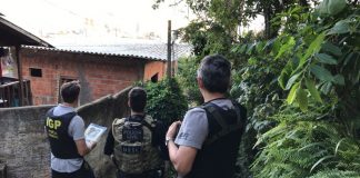 Três policiais vistos de costas usando coletes à prova de bala com escritos da polícia civil em frente à casas de favela