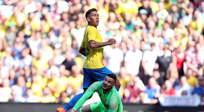 jogador firmino olha para fora da imagem e o goleiro adversário na mesma direção, caído no chão; grande torcida ao fundo