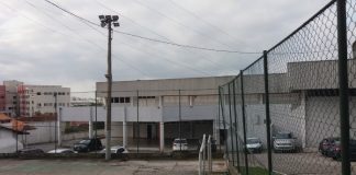 Vista geral do clube, com quadra de cimento em primeiro plano, cercada; atrás o prédio principal, com alguns carros estacionados na frente