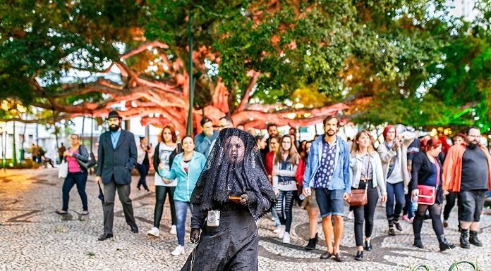 mulher trajada de uma típica viúva anda à frente de um grupo de pessoas na praça 15