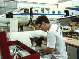 rapaz usando equipamento de segurança do trabalho mexe em peça de aviação com ferramentas em um hangar; há uma aeronave no fundo