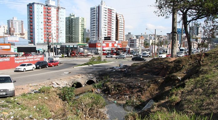 rua entre as cidades onde há um esgoto a céu aberto e muito lixo em volta no canteiro; alguns carros passando e ao fundo os prédios da cidade de são josé