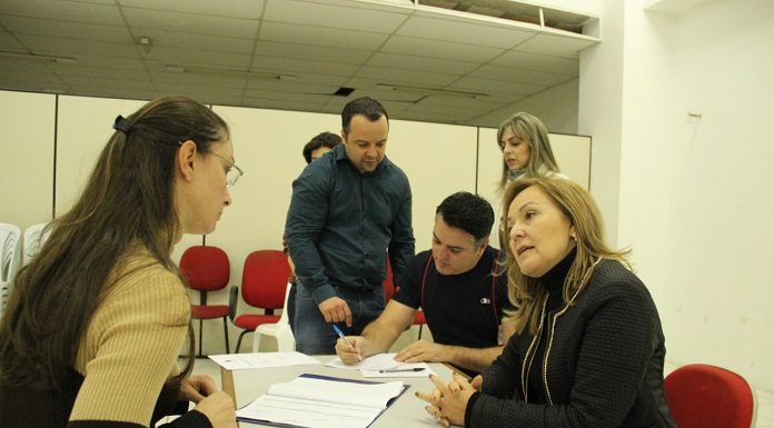 duas mulheres conversam de cada lado de uma mesa; há outras pessoas em pé ao fundo conferindo documentos sobre a mesa