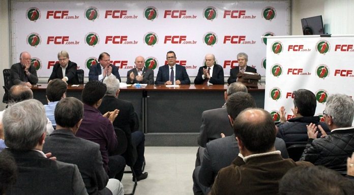 pequena sala de auditório com pessoas sentadas vistas de costas e ao fundo, na bancada, o presidente eleito ao centro de outras pessoas; todas são homens