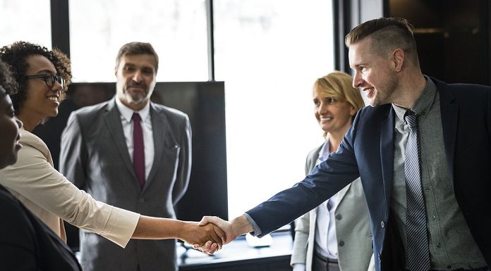 dois jovens se cumprimentam sobre uma mesa de trabalho com outras pessoas olhando em volta