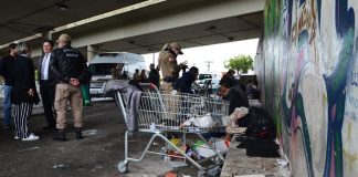 policial feminina conversa olhando no celular com um morador de rua sentado no meio fio ao longo de um muro sob o viaduto; em primeiro plano há um carrinho de supermercado; ao lado algumas outras pessoas (policias e agentes públicos de saúde) conversam