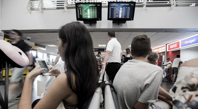 homem e mulher sentados em cada lado de bancos de espera no saguão do aeroporto; há dois monitores em uma parede no alto ao fundo