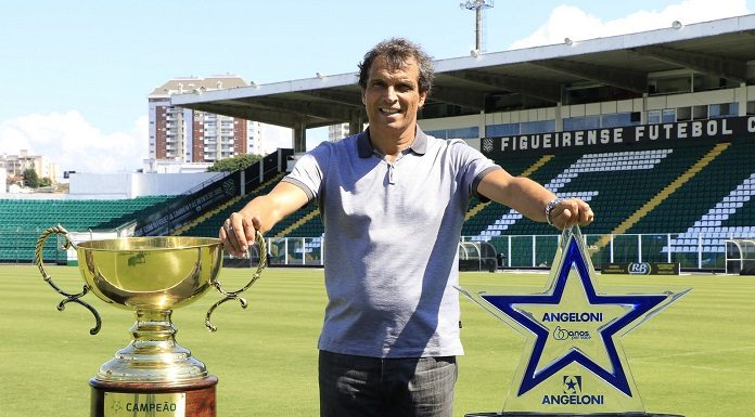 técnico posa sorridente para foto com as mão sobre dois troféus bem ao centro do gramado do estádio do figueirense; arquibancadas ao fundo