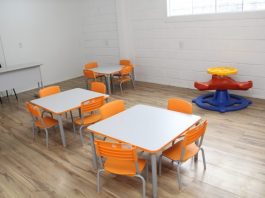 sala de aula com duas mesas infantis cercadas por cadeiras laranjas para crianças e alguns brinquedos