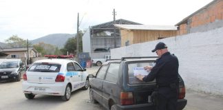 agente de trânsito visto de costas colocando um adesivo da prefeitura em um fiat uno abandonado em uma rua, com um veículo gol da prefeitura estacionado ao lado