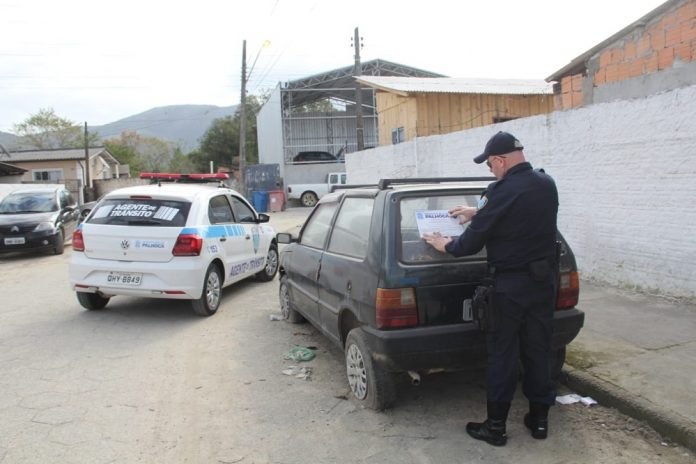 agente de trânsito visto de costas colocando um adesivo da prefeitura em um fiat uno abandonado em uma rua, com um veículo gol da prefeitura estacionado ao lado