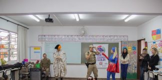 em uma sala de aula, crianças pequenas sentadas no chão assistem a apresentação de uma mulher vestida de super mulher e um policial militar de farda; ha outras professoras de pé assistindo; a maioria está sorrindo