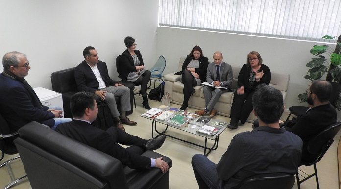 grupo de homens e mulheres em reunião em uma sala com sofás e poltronas em volta de uma pequena mesa de centro ao lado de uma janela