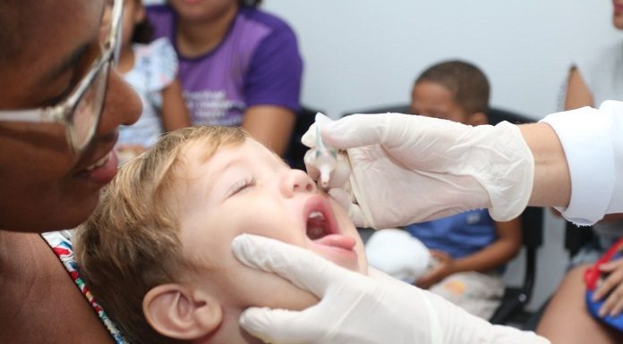 criança com os fechados e boca aberta pra cima coloca a língua pra fora ao receber gotas de vacina seguradas por mãos usando luvas; uma mulher segura a criança