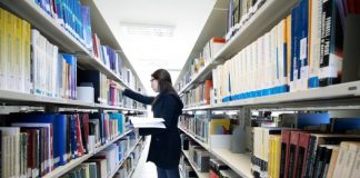 mulher confere prateleira em um corredor no meio de estantes de livros em uma biblioteca