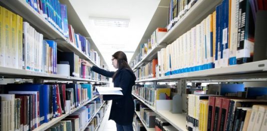 mulher confere prateleira em um corredor no meio de estantes de livros em uma biblioteca