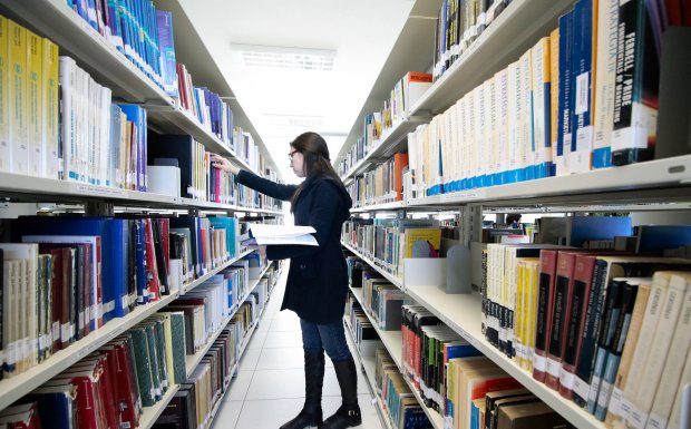 mulher confere prateleira em um corredor no meio de estantes de livros em uma biblioteca