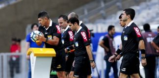 quatro árbitros com uniforme com propaganda na manga entram em campo juntos; o juiz pega a bola de cima de um pedestal no campo