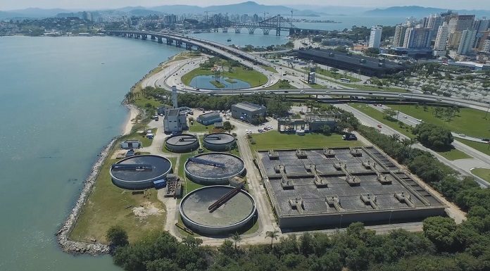 imagem aérea de uma estação de tratamento de esgoto em área na beira do mar no centro de florianópolis, onde ao fundo é possível ver prédios e as pontes em dia de sol