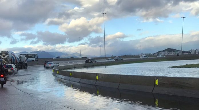 foto tirada de dentro de um carro mostra uma curva fechada, onde há um lago ao lado, com água na pista e alguns carros passando