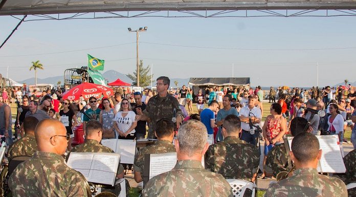 homens com roupa camuflada do exército tocando em uma orquestra; maestro em pé de frente para câmera rege a música e populares ao fundo observam