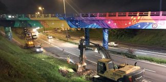 foto tirada de cima de um barranco mostra uma retroescavadeira abrindo buraco ao lado da rodovia; outros operários e máquinas circulam pelo acostamento, sob um viaduto colorido