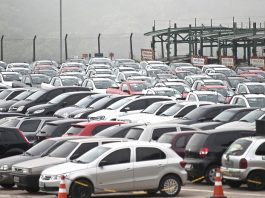 pátio lotado e carros estacionados de diversos modelos; com um caminhão cegonha ao fundo