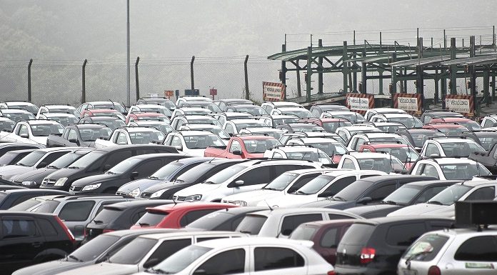 pátio lotado e carros estacionados de diversos modelos; com um caminhão cegonha ao fundo